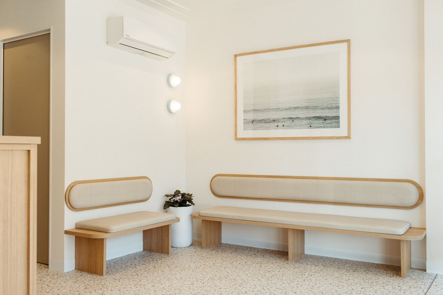 Minimalist waiting room with two wooden benches, white walls, round sconces, and a framed coastal artwork above.