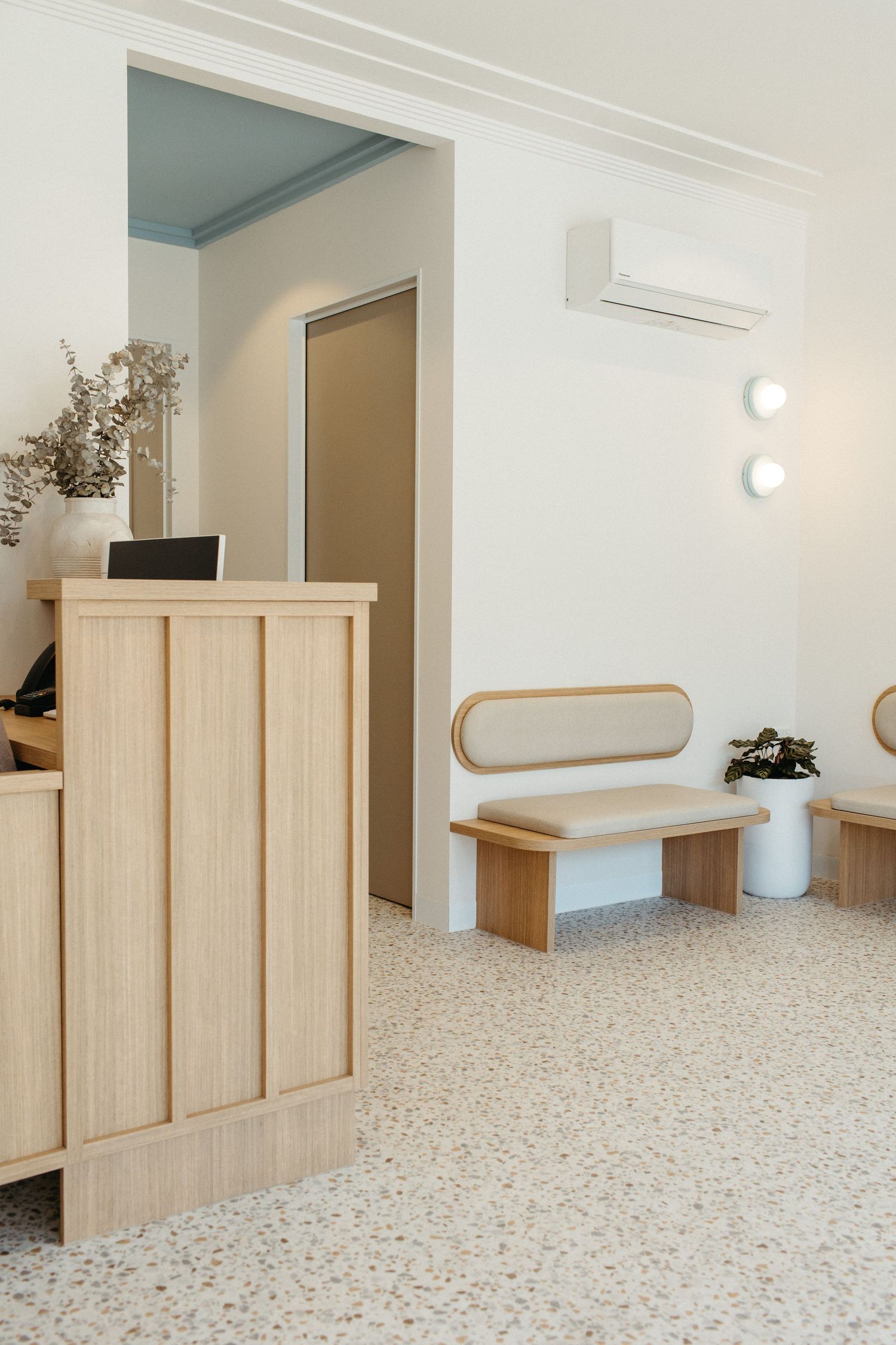 Modern minimalist reception area with light wood desk, terrazzo floors, bench seating, and wall-mounted AC unit.
