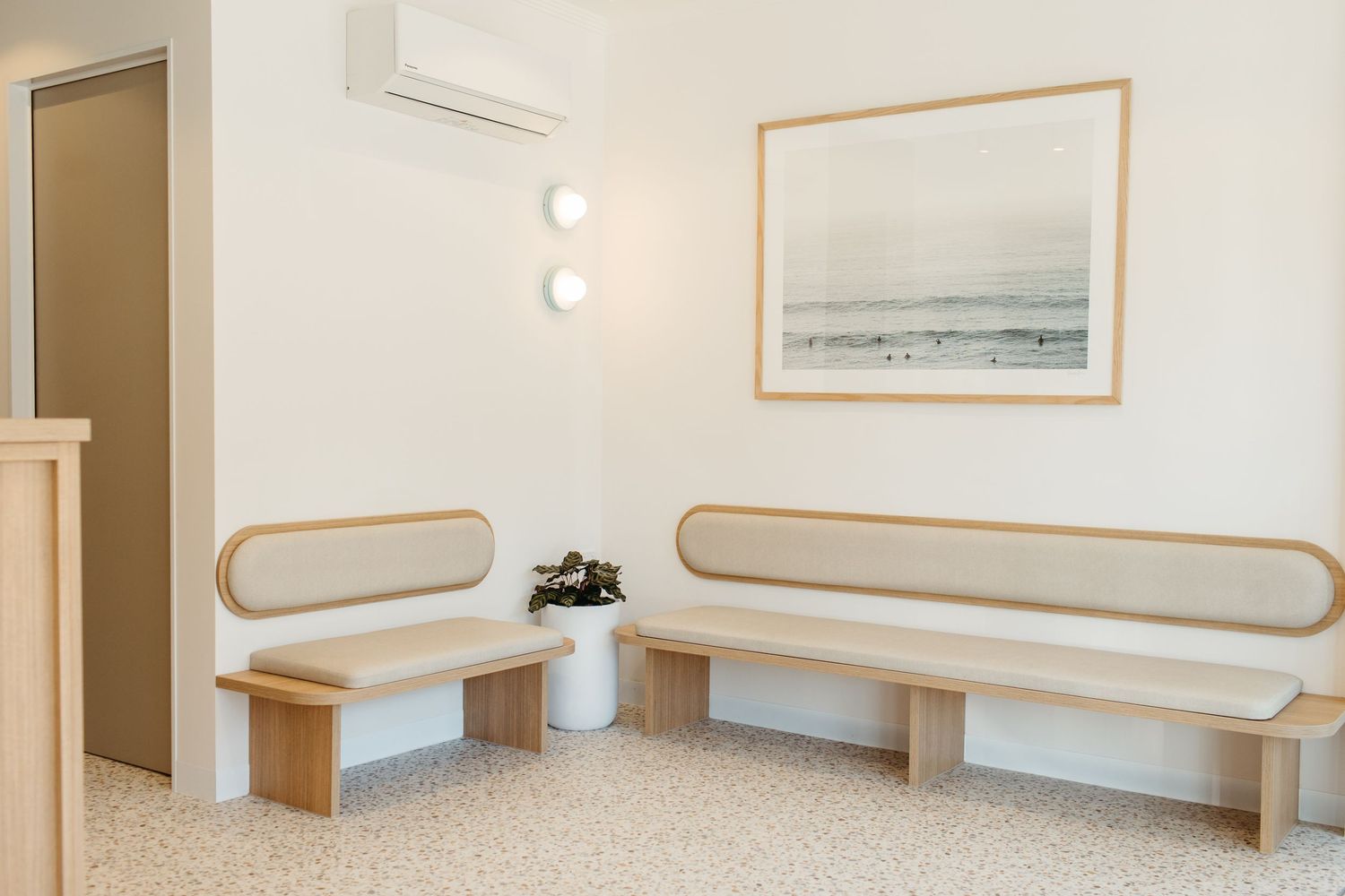 Minimalist waiting room with two wooden benches, soft lighting, wall art, and terrazzo floors in a modern interior.