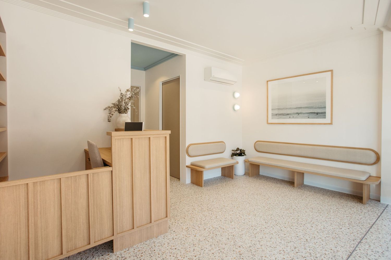 Modern minimalist waiting room with light wood benches, terrazzo floors, and a framed coastal artwork on white walls.