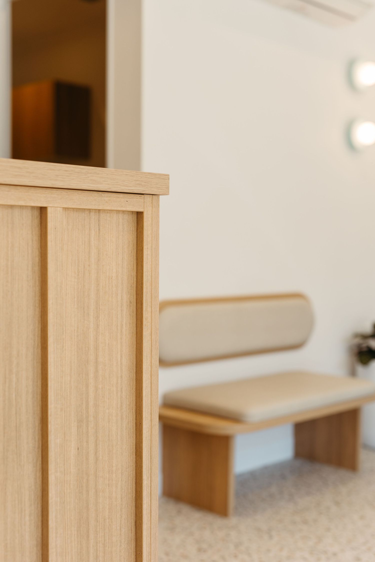 Minimalist waiting area with light wood bench, beige cushion, and wooden partition on terrazzo floor.