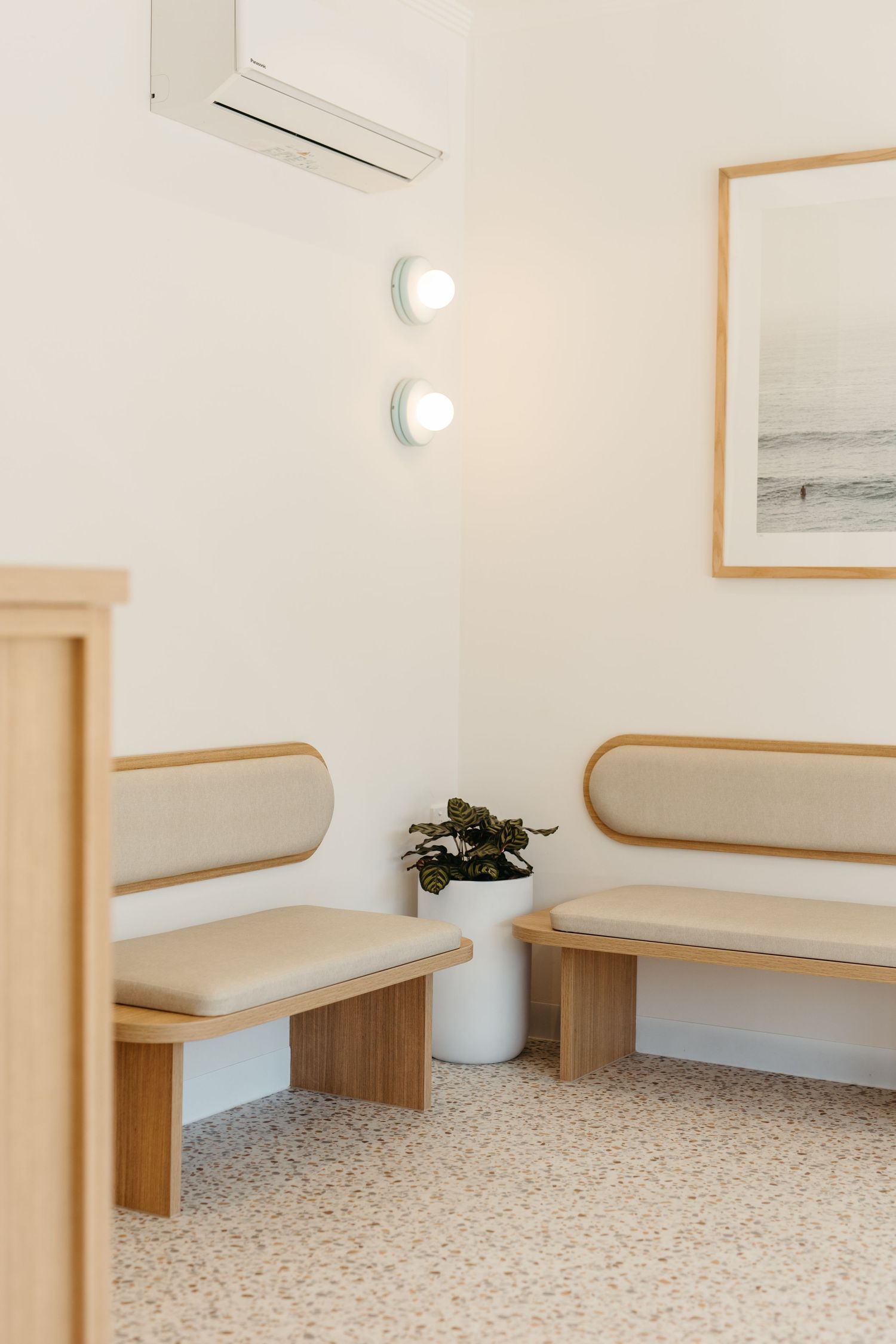 Minimalist waiting room with two wooden benches, cream upholstery, terrazzo floor, wall sconces, and a framed coastal print.