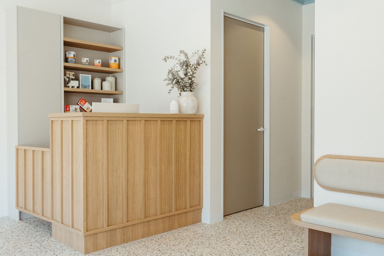 Modern reception desk in light oak wood with built-in shelving, terrazzo floors, and a neutral door in a minimalist interior.