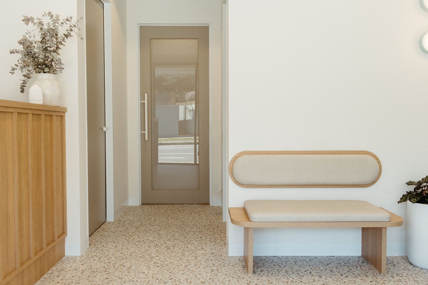 Modern minimalist entryway with a wooden bench, terrazzo flooring, taupe door, and neutral white walls.