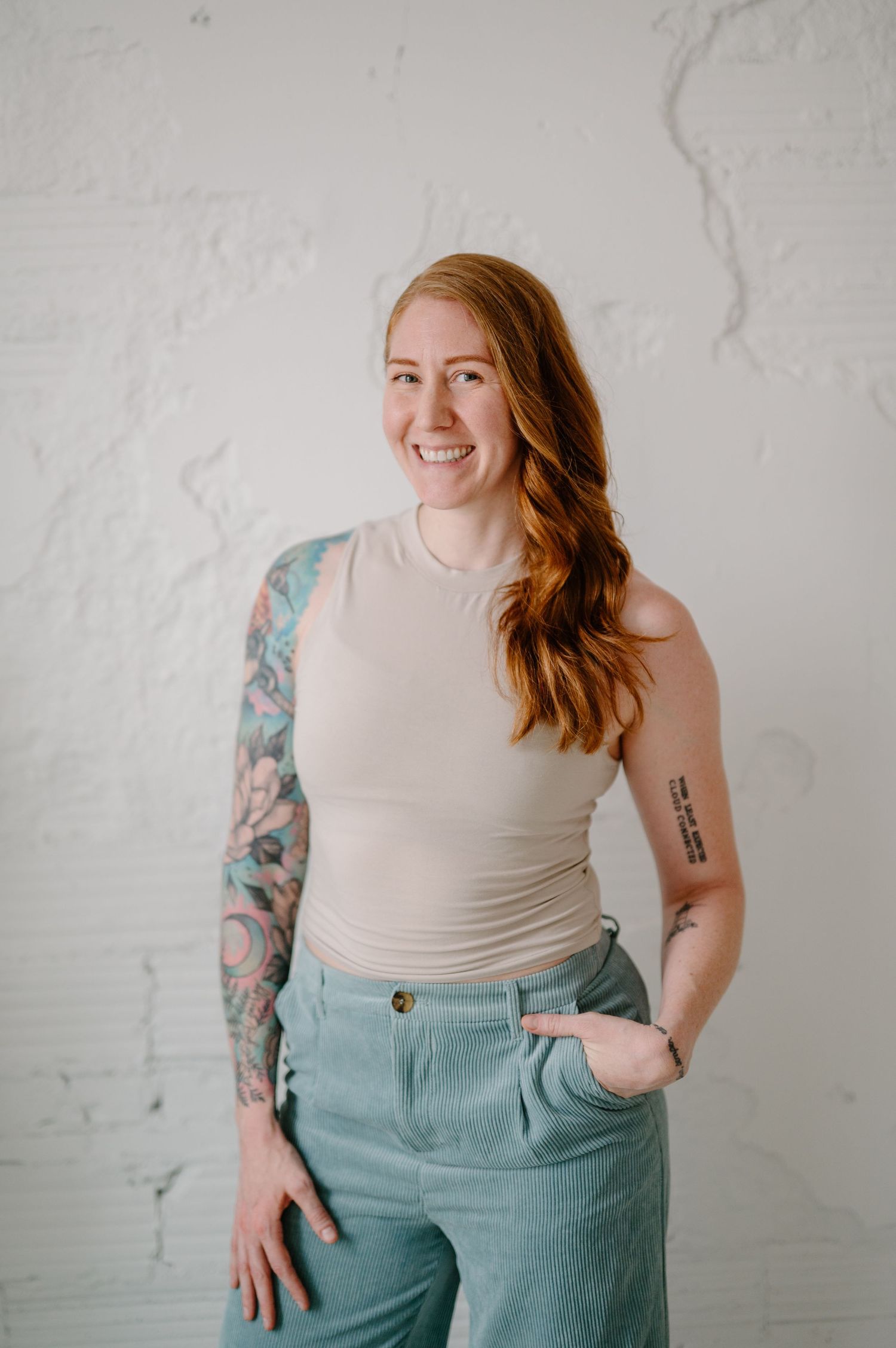 Smiling redhead woman with sleeve tattoo wearing beige tank top and mint green corduroy pants against white brick wall.
