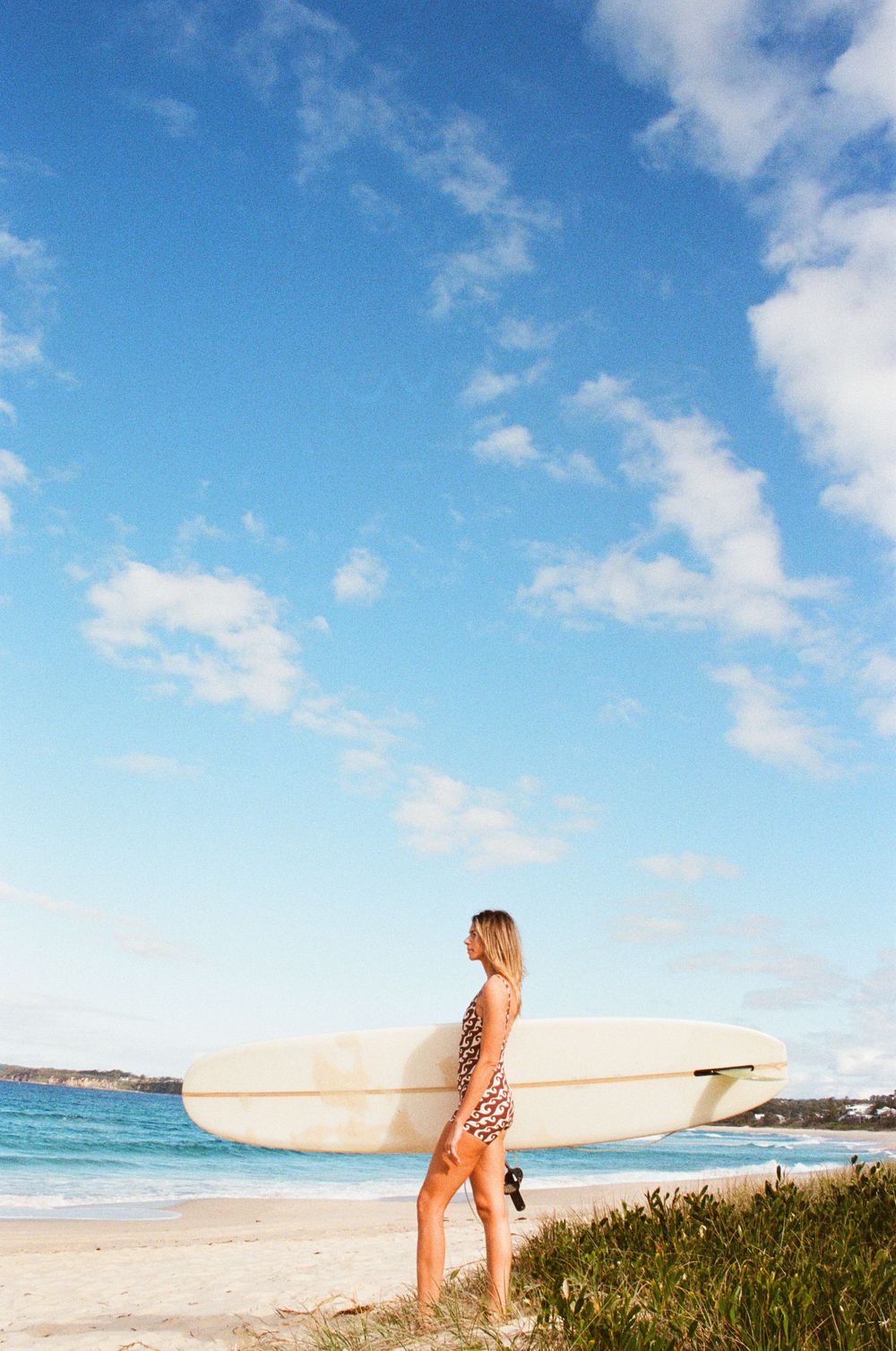 Woman in leopard print swimsuit holding a white longboard surfboard on a sunny beach with blue sky and ocean backdrop.