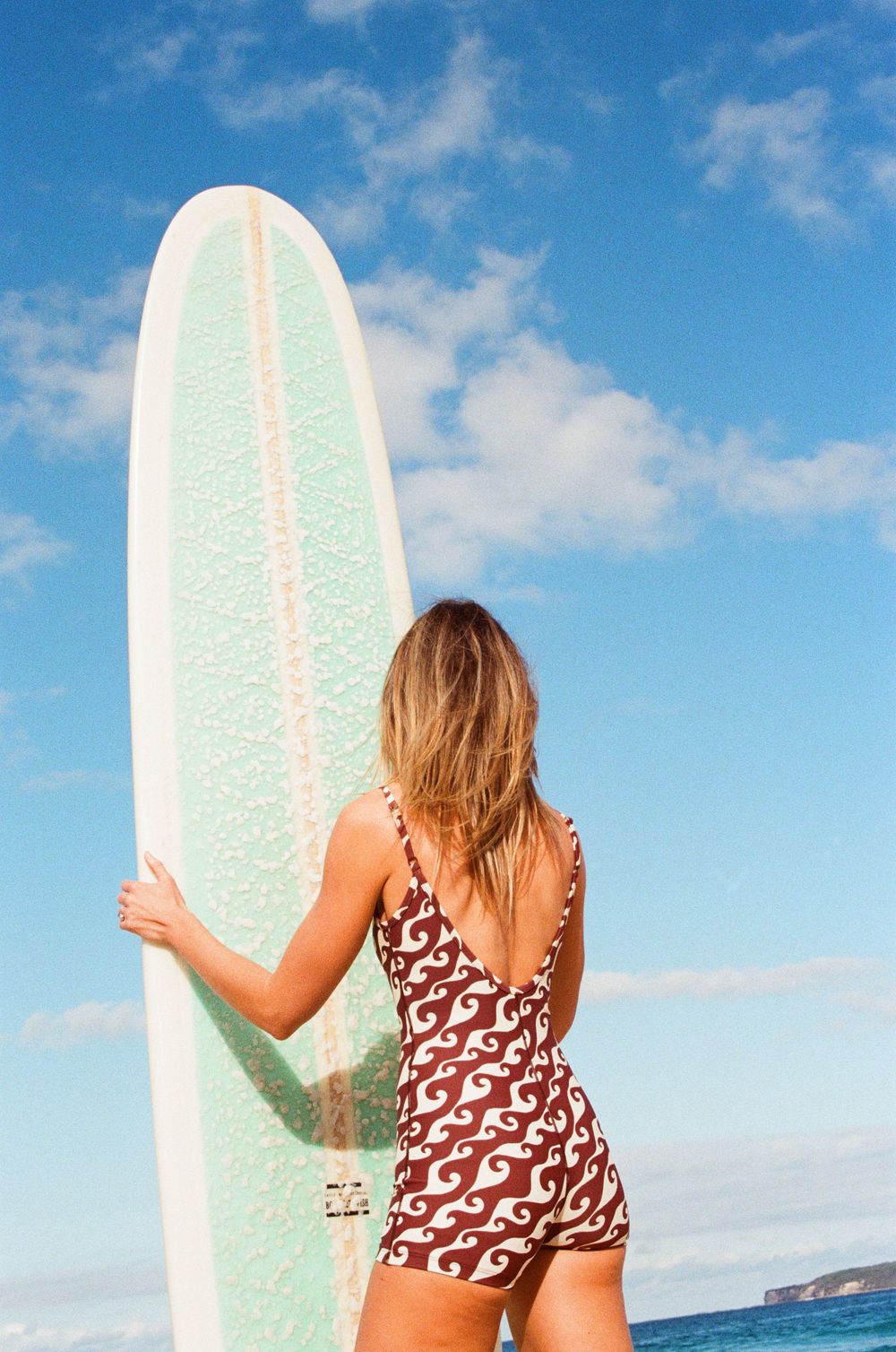 Woman in patterned swimsuit holding a white longboard surfboard on a sunny beach with blue sky.