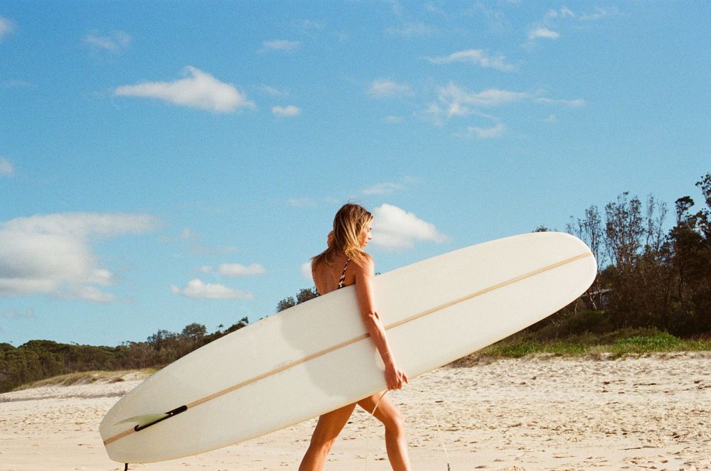 Woman carrying a white longboard surfboard on a sunny sandy beach under a blue sky.