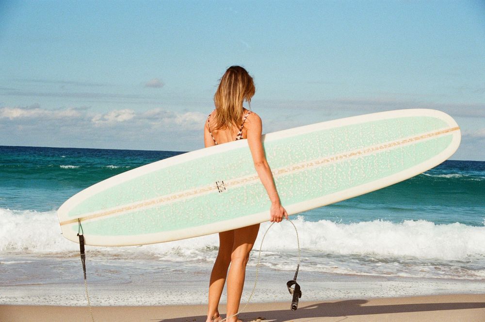 Blonde woman carrying a mint green longboard surfboard walking toward ocean waves on a sunny beach.