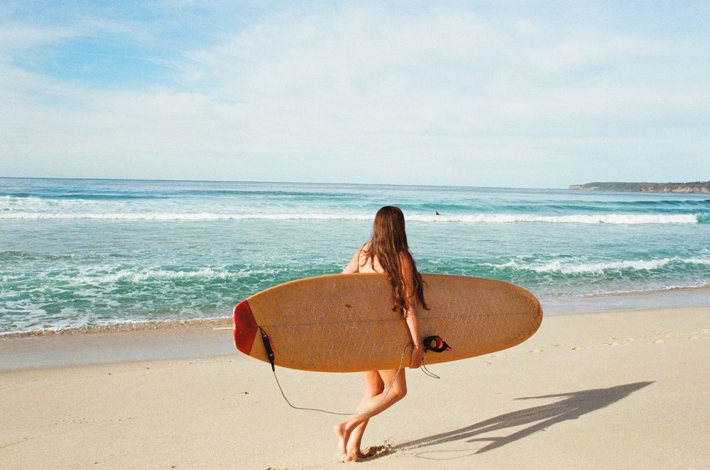 Woman in bikini carrying a wooden surfboard walking toward ocean waves on a sunny sandy beach.