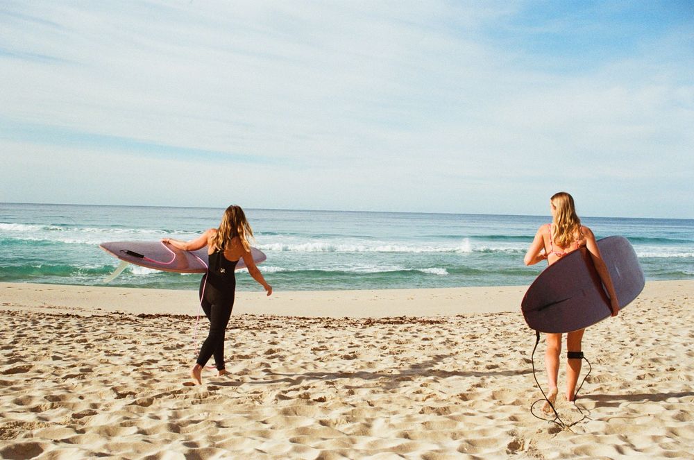 Two women carrying surfboards walk toward ocean waves on a sunny sandy beach.