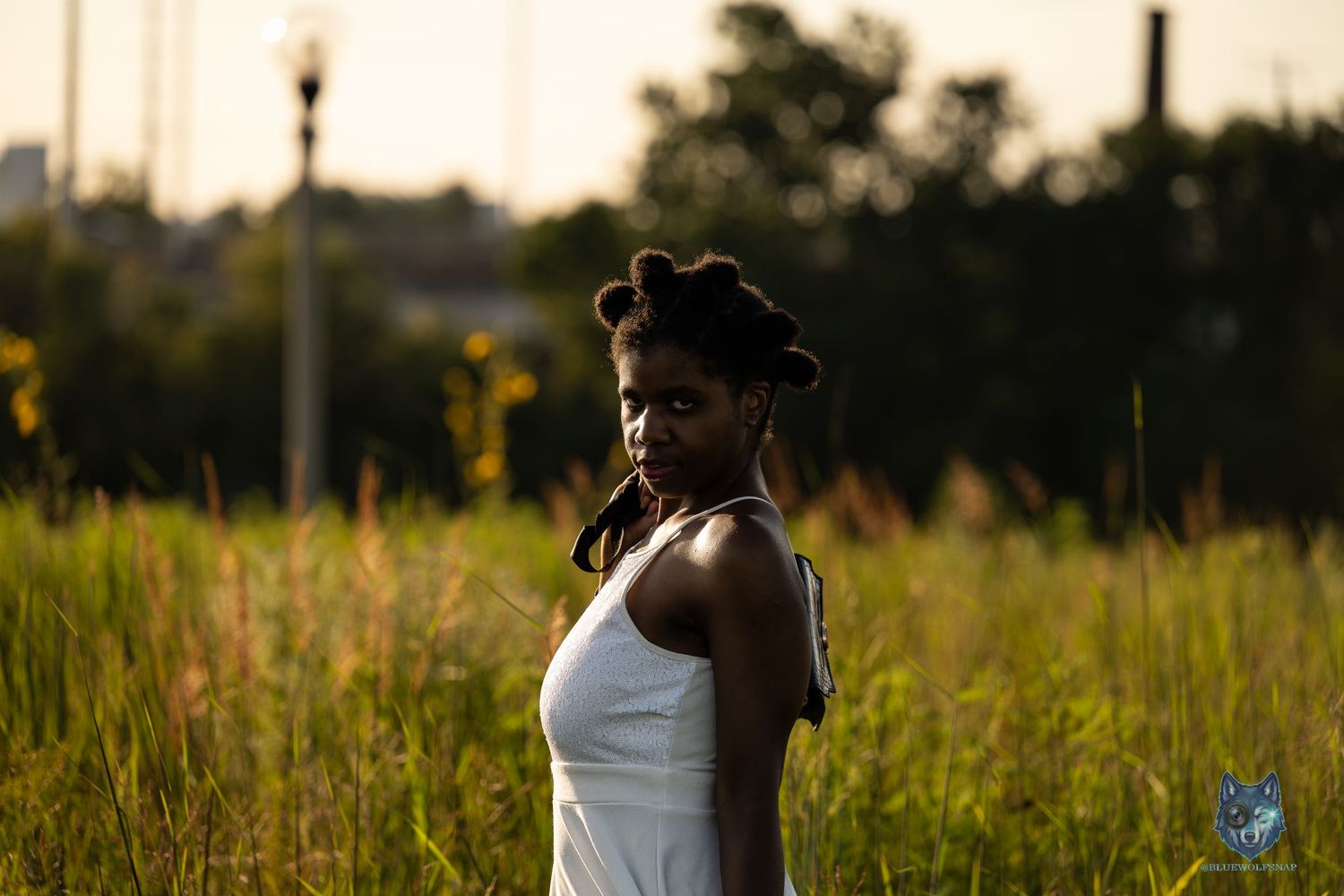 Young Black woman in white dress standing in tall green grass field during golden hour sunset.