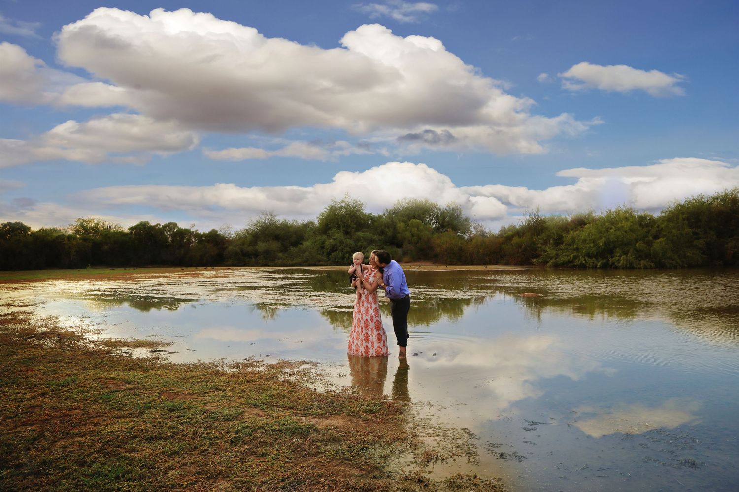 A couple in an embrace stands in shallow water, their reflections mirrored on the surface, under a blue sky with puffy white clouds.