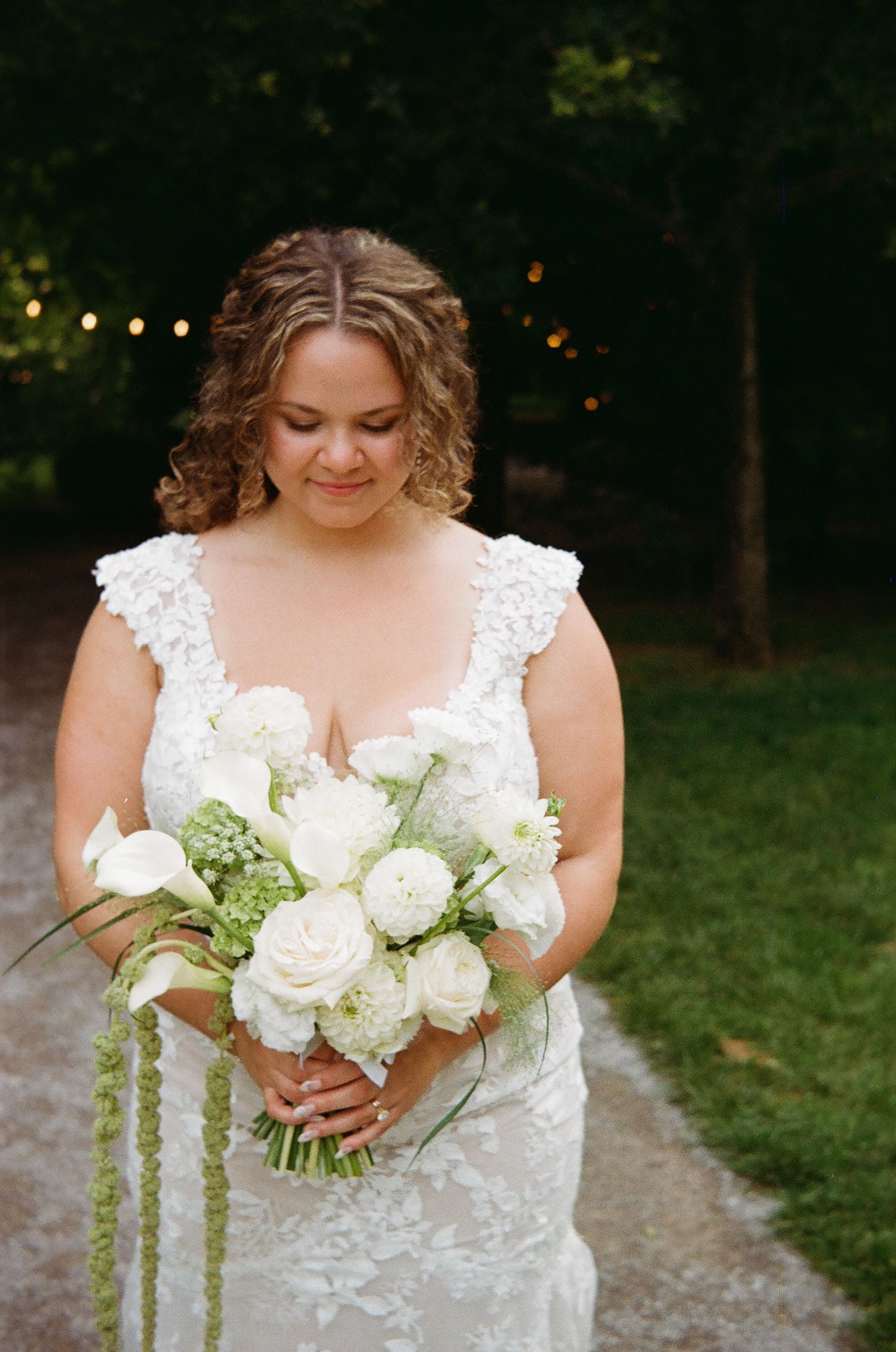 Bride in lace wedding dress holding white rose bouquet with greenery, looking down outdoors at dusk. Cranberry Creek Gardens Ontario