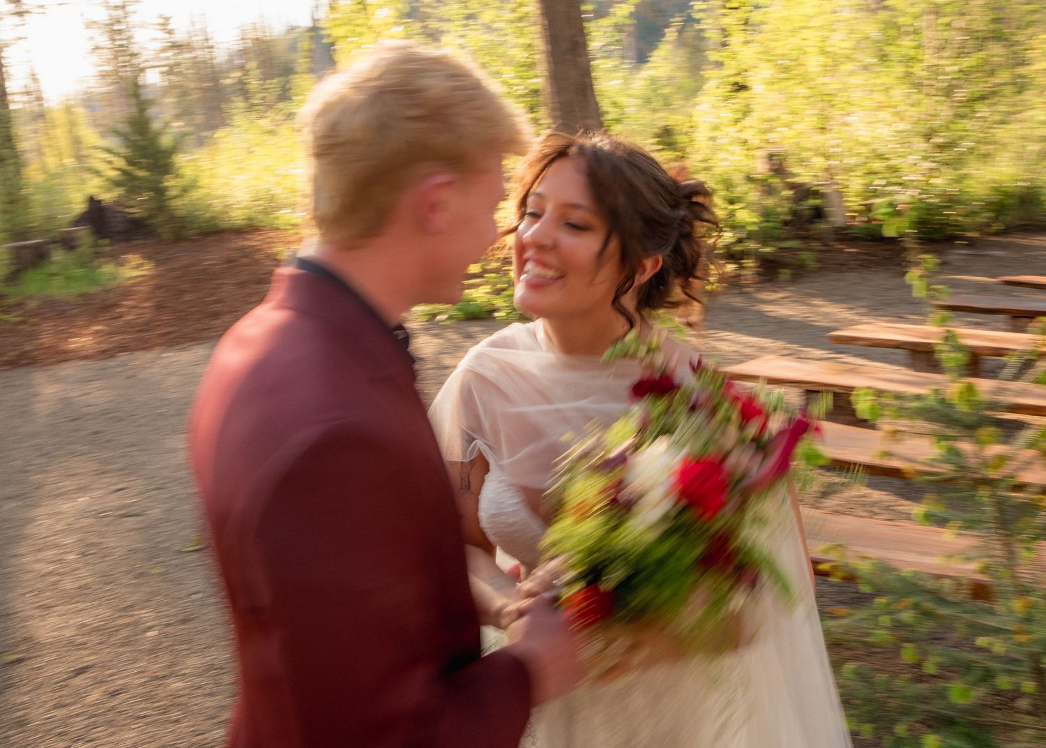 A bride holding red roses smiles at a blond groom in a burgundy suit outdoors in a forest wedding setting.
