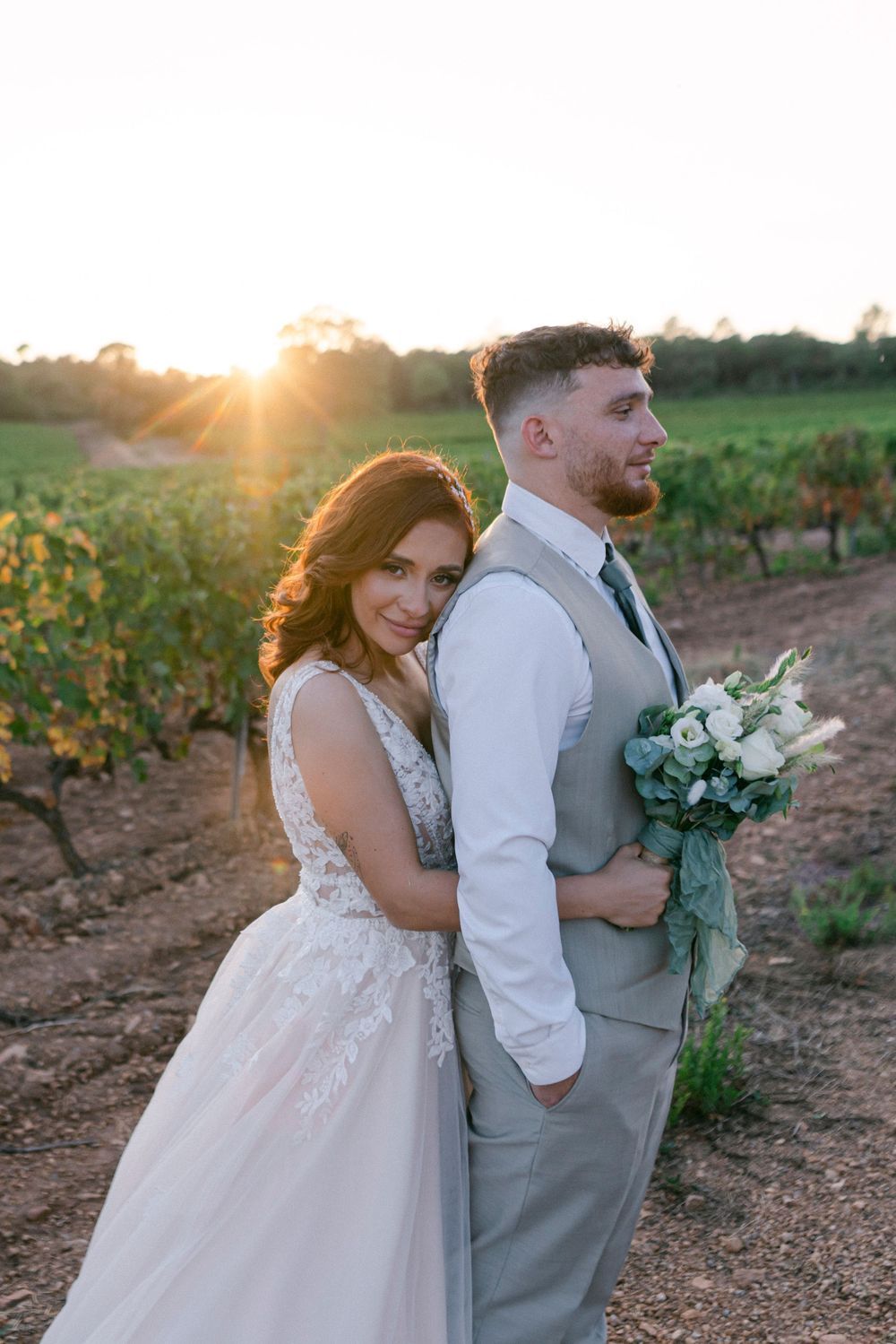 Bride and groom pose back-to-back in a vineyard at golden sunset, bride holding white bouquet in lace wedding gown.