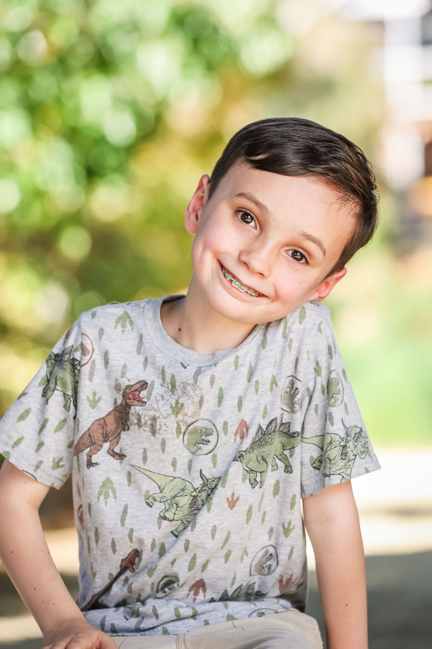 Smiling boy with braces wearing a dinosaur print t-shirt outdoors with green foliage background.