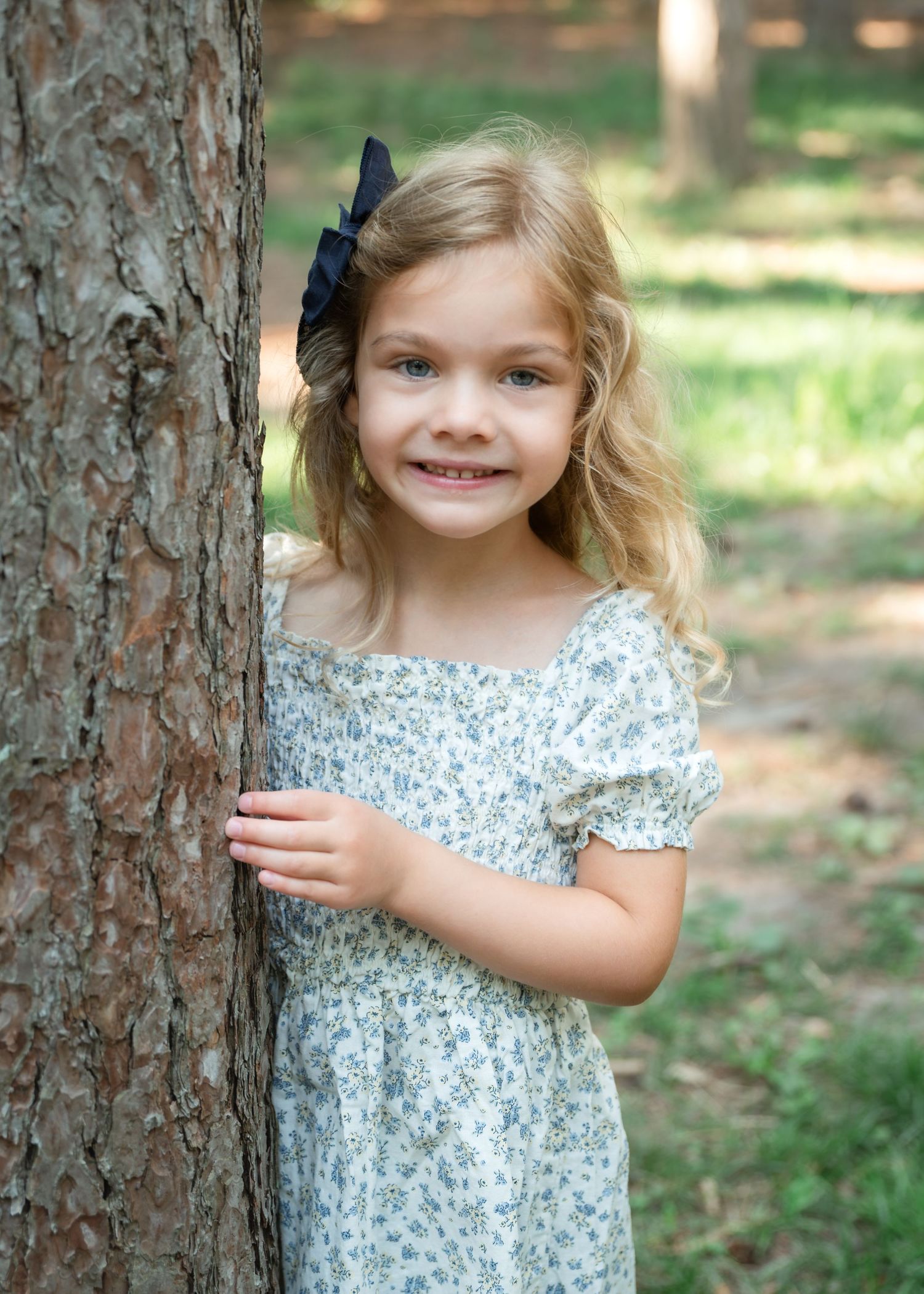 Young blonde girl in floral dress smiling beside a tree in a sunlit park, wearing a navy bow hair clip.