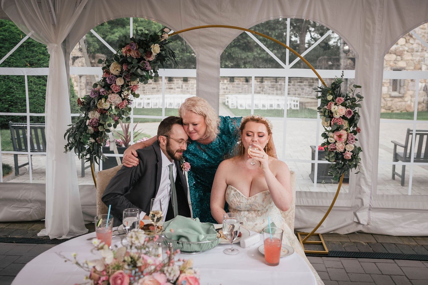 Bride's mother embracing the groom and resting against the bride's shoulder during a quiet moment at the sweetheart table.