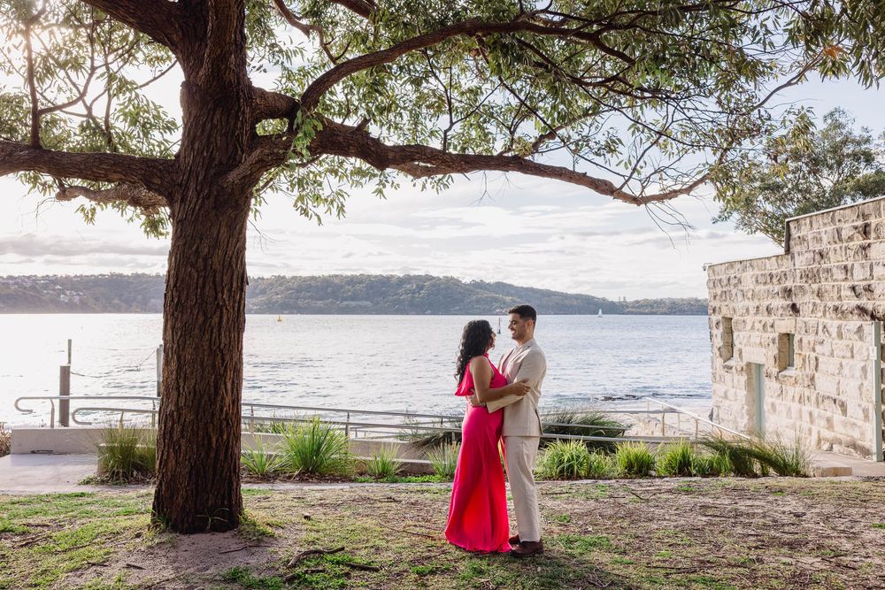Couple in formal wear embracing under a tree by Sydney Harbour waterfront at sunset.
