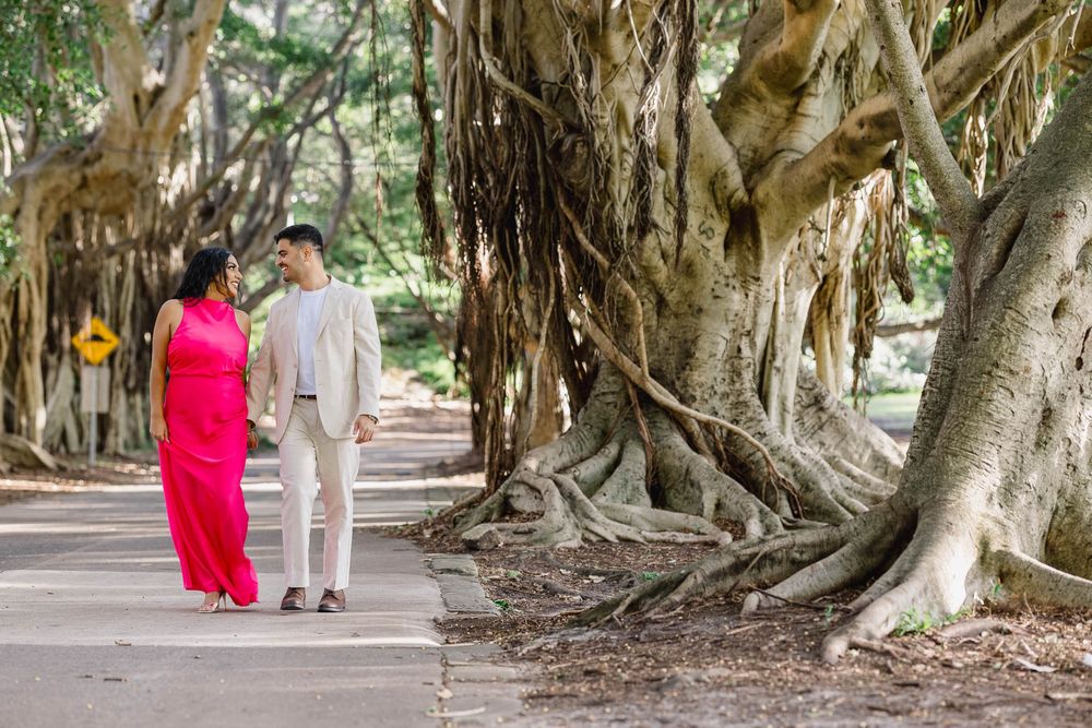 Couple holding hands walking along a tree-lined path surrounded by large banyan trees in a lush park.