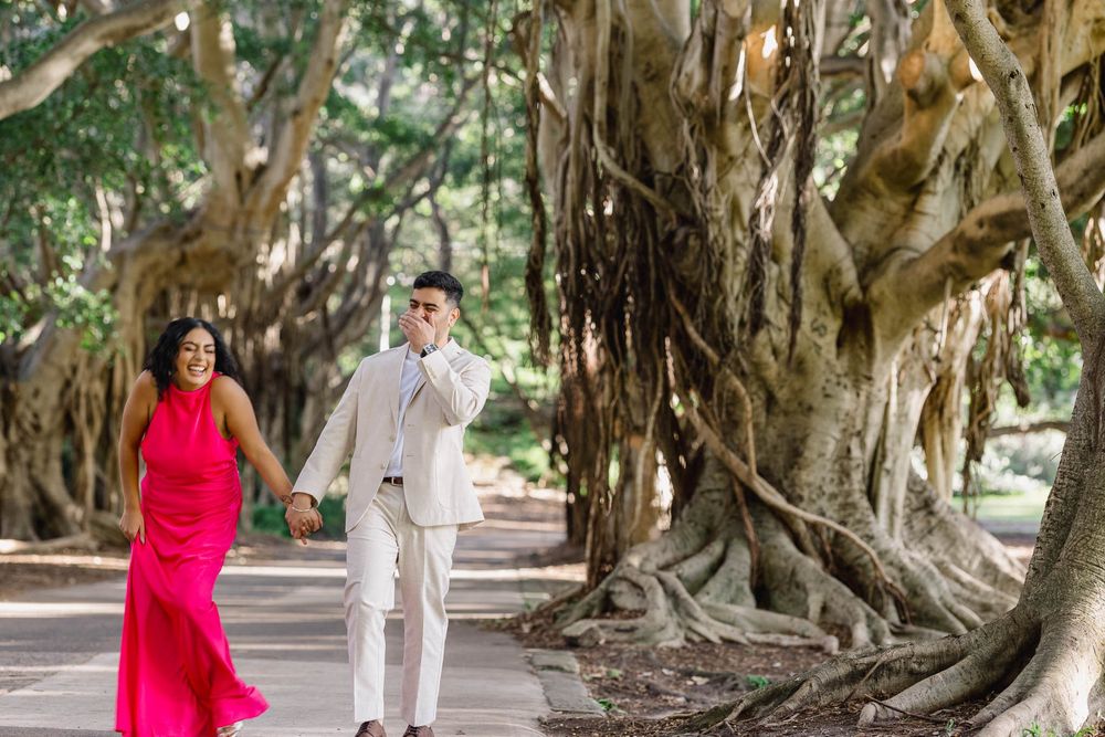 A couple holds hands walking past large banyan trees, woman in pink dress, man in white suit.