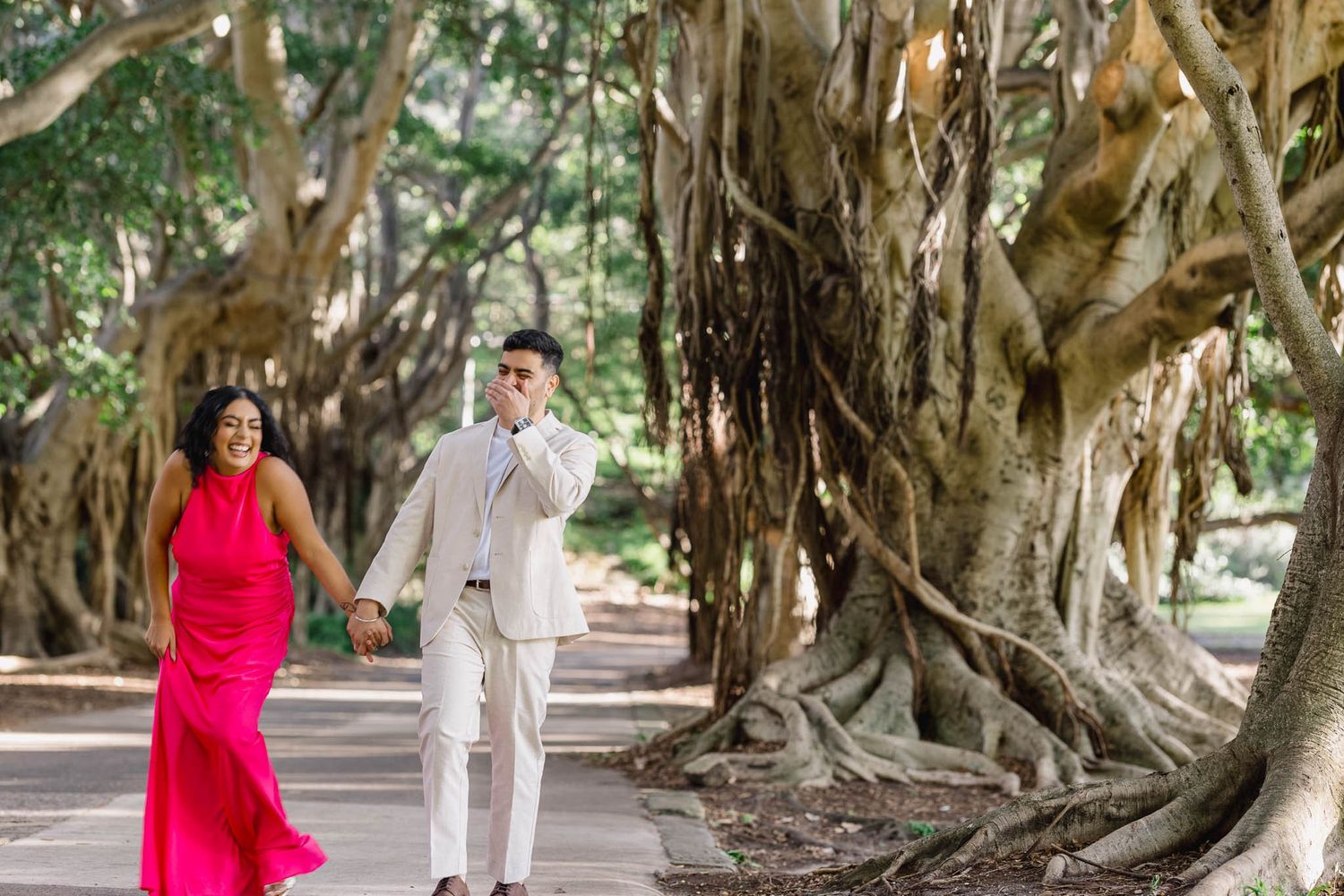 A couple holds hands walking past large banyan trees, woman in pink dress, man in white suit.