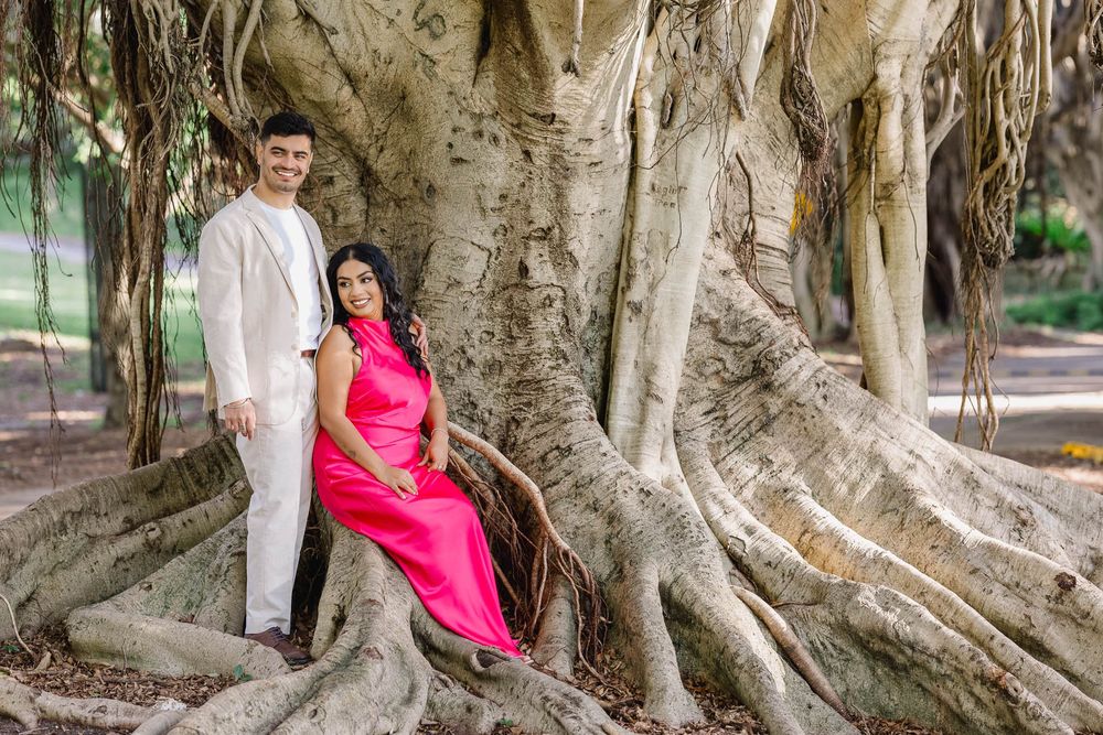 Couple posing in front of large banyan tree roots, woman in pink dress, man in white suit, outdoor park setting.