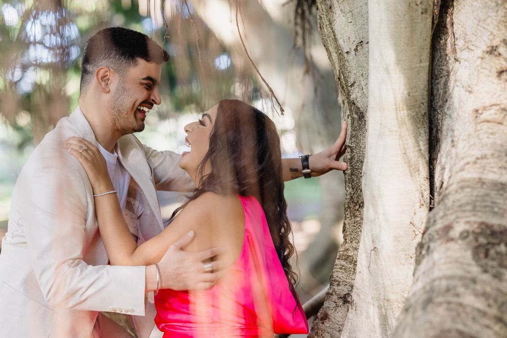 Couple laughing and embracing by a large tree, woman in pink dress, man in beige suit, outdoor engagement photo.