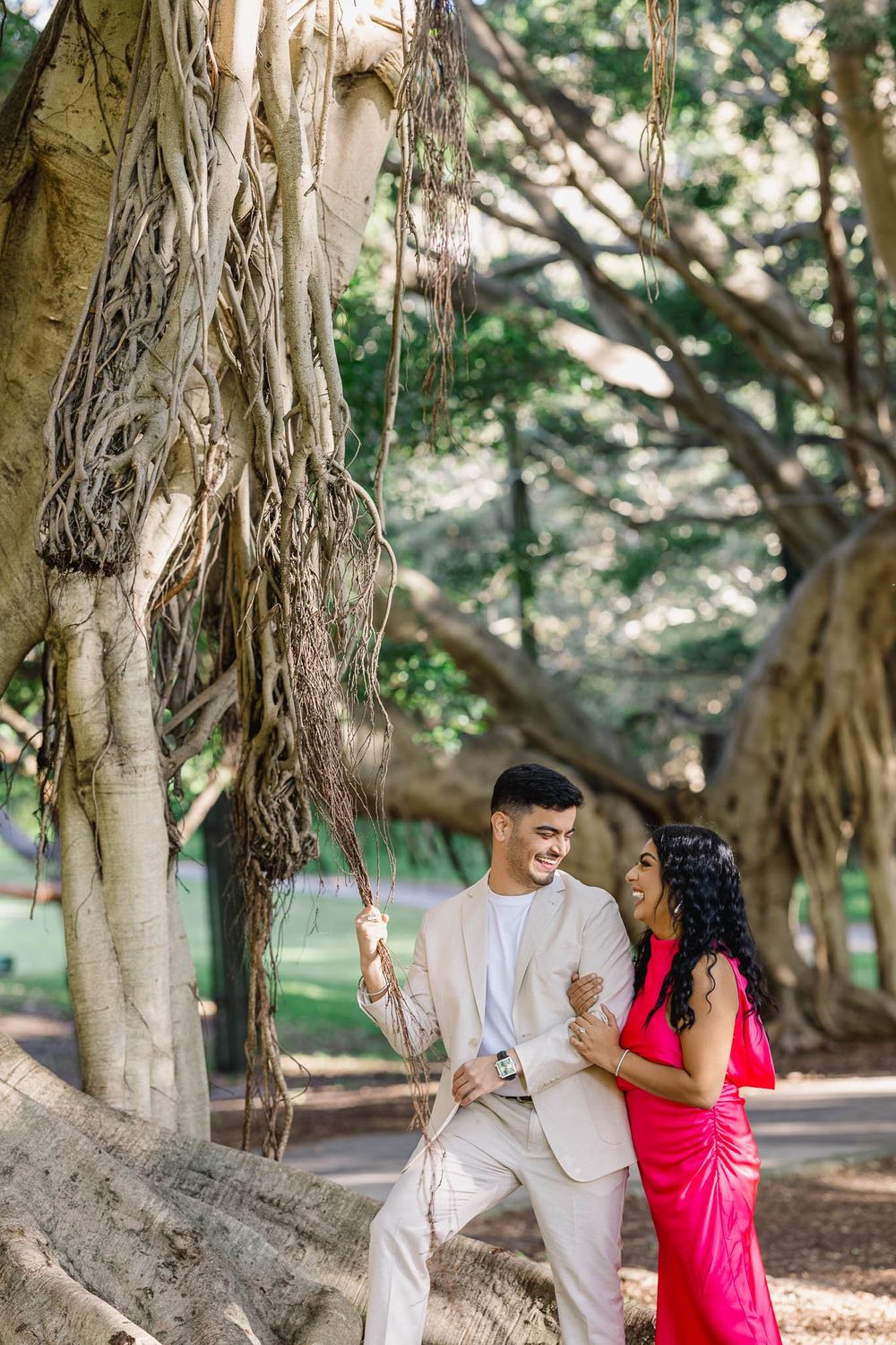 Couple laughing together near banyan tree roots in a lush park, man in beige suit, woman in pink dress.