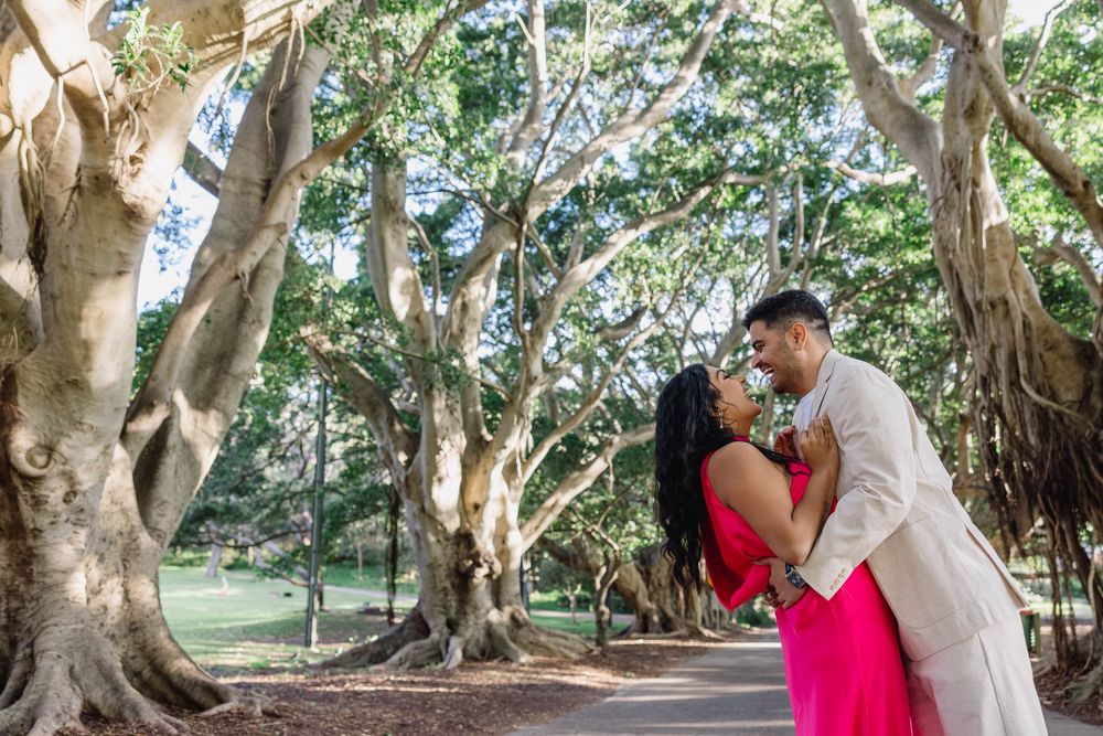 Romantic couple sharing a kiss under sprawling banyan trees in a sunlit park, woman in pink dress.