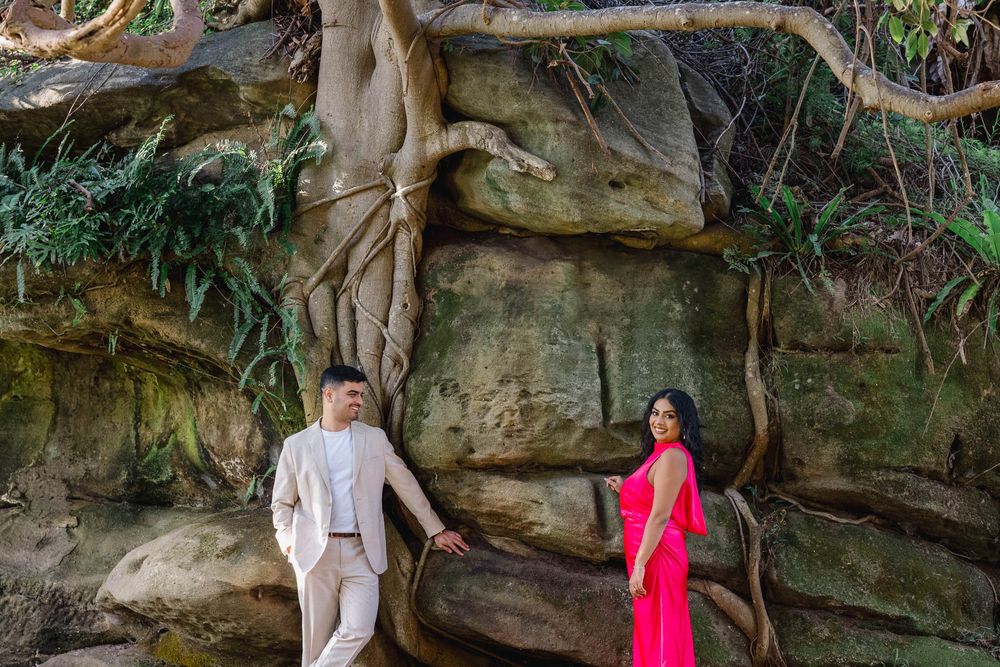 Couple posing by large rock formation with tree roots, man in beige suit and woman in pink dress.