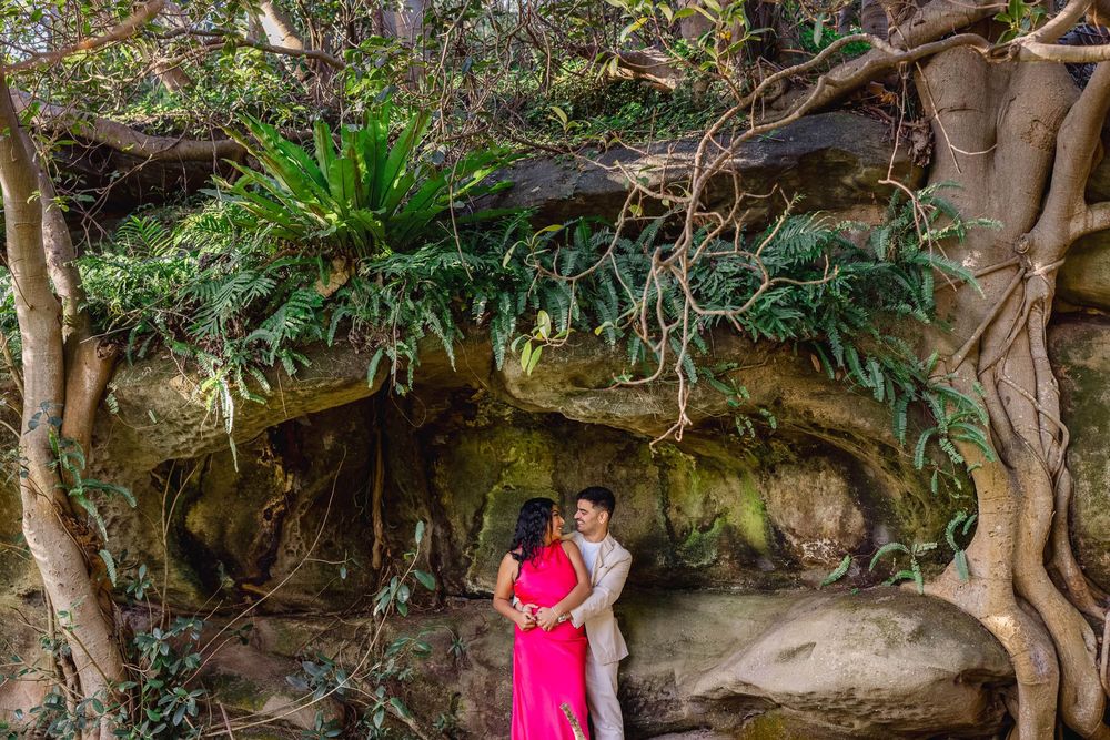 Couple in pink and beige attire embracing under ancient fig trees with lush greenery and mossy rocks.