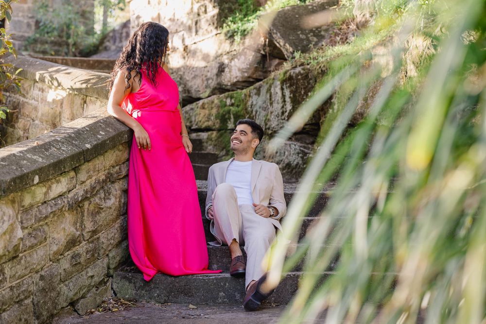 Couple posing outdoors on stone steps, woman in pink dress, man in beige suit, surrounded by lush greenery.