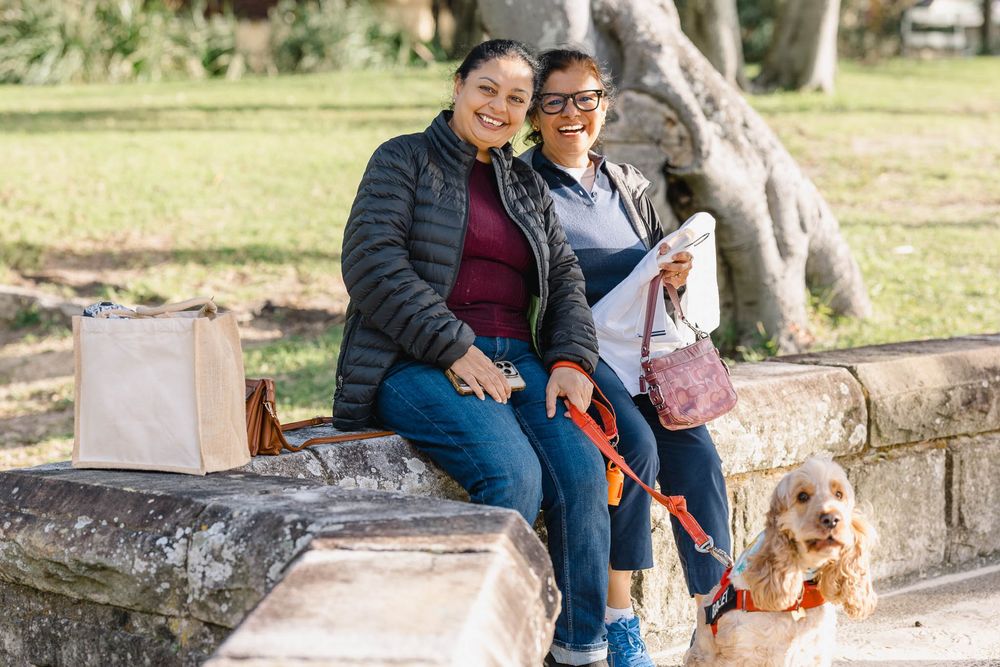 Two women sitting on a stone bench in a park with a golden cocker spaniel dog, smiling outdoors.