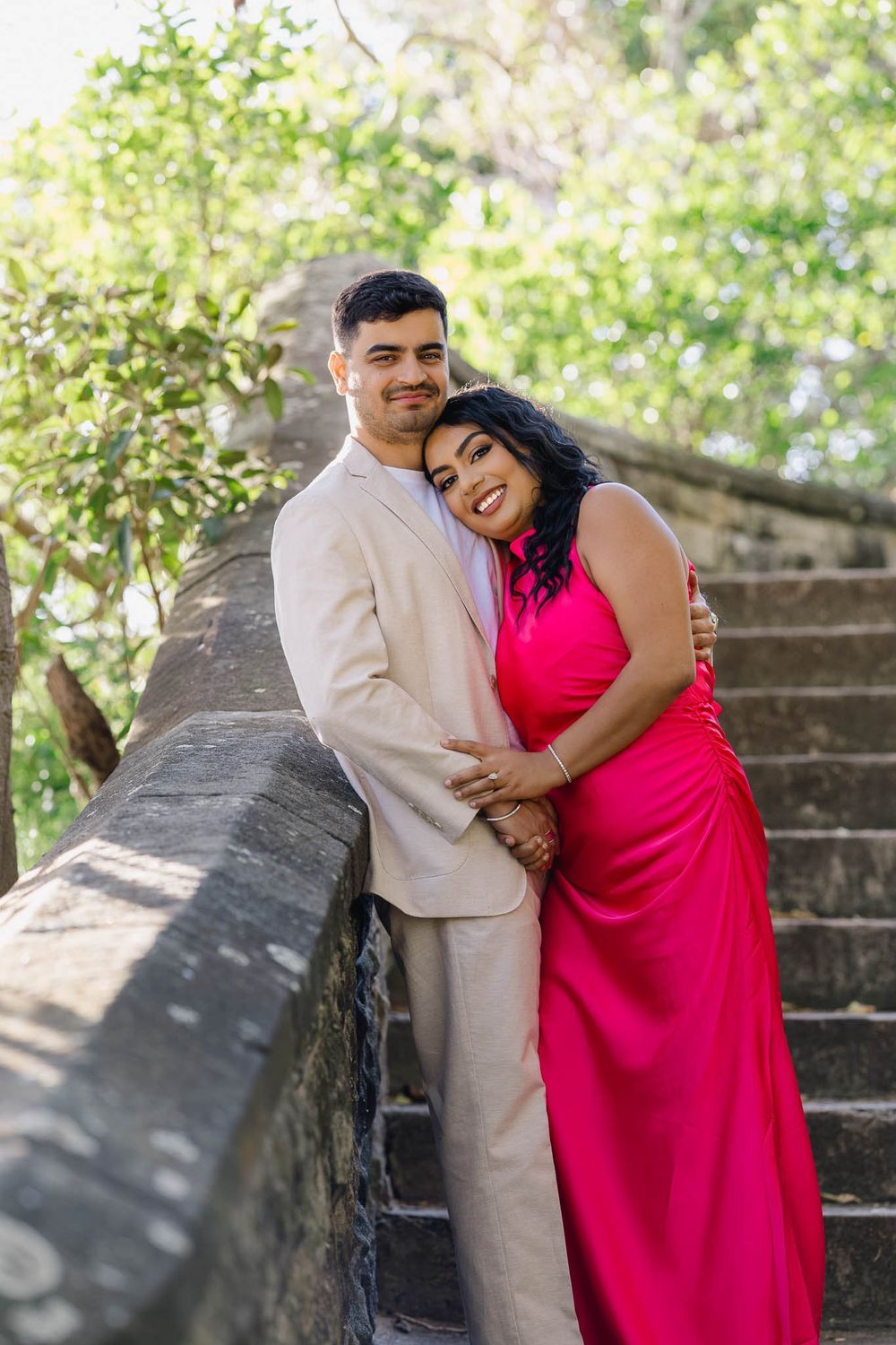 Couple posing on stone steps, man in beige suit, woman in pink dress, embracing outdoors.