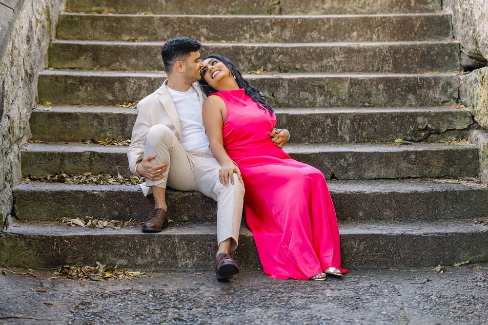 Couple in formal wear sitting on stone steps, woman in pink gown leaning on man in white suit.