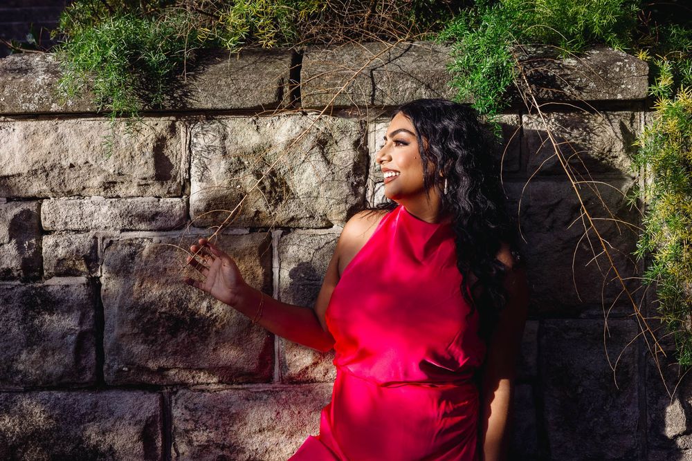 Woman in red dress leaning against stone wall with green foliage in sunlight.