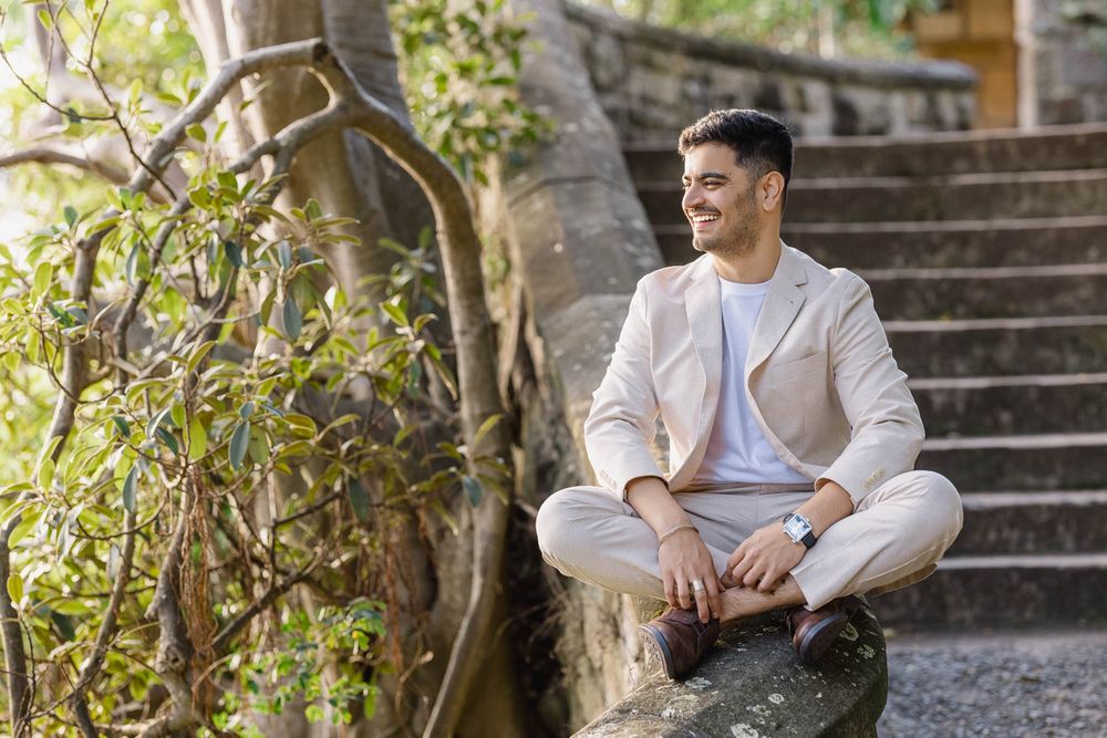 Man in beige suit sitting cross-legged on stone wall, smiling outdoors near trees and stone steps.