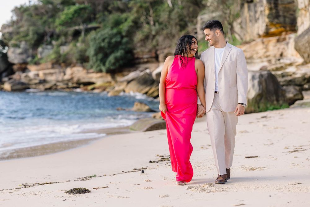 Couple walking hand in hand on a sandy beach, woman in red dress, man in beige suit, rocky cliffs behind.