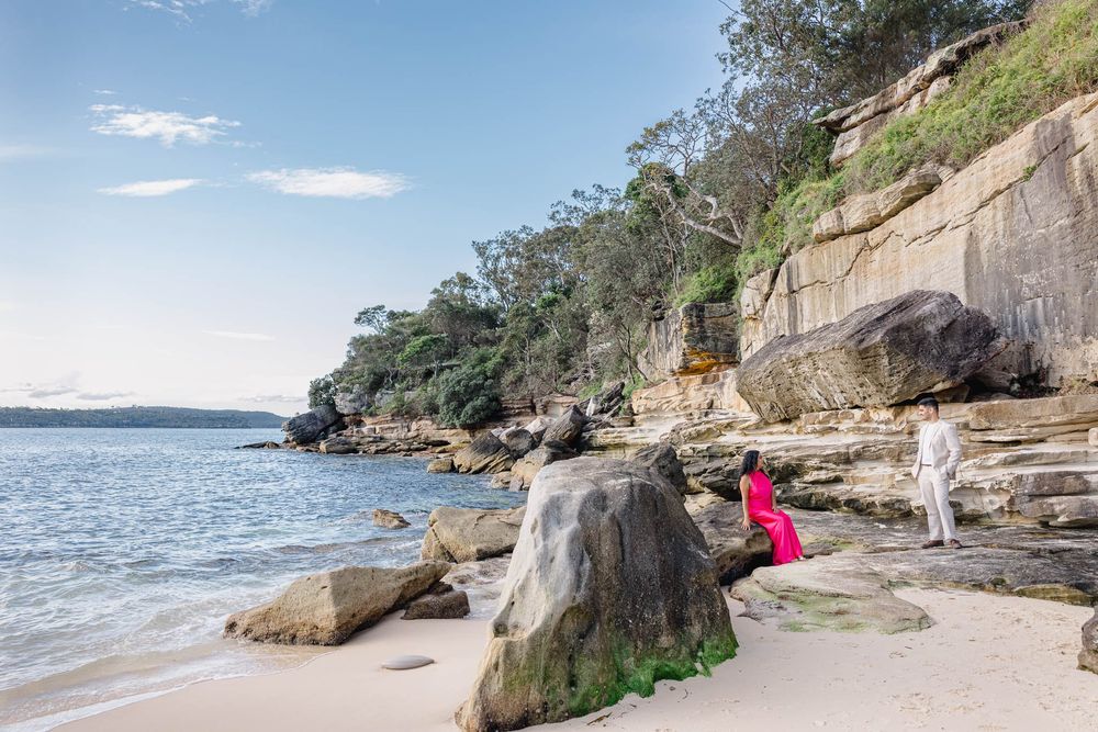 Couple in pink and white outfits on a rocky sandy beach with sandstone cliffs and calm blue water.