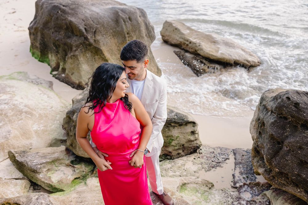 Couple in romantic embrace on rocky beach, woman in red dress, man in white shirt, ocean waves in background.