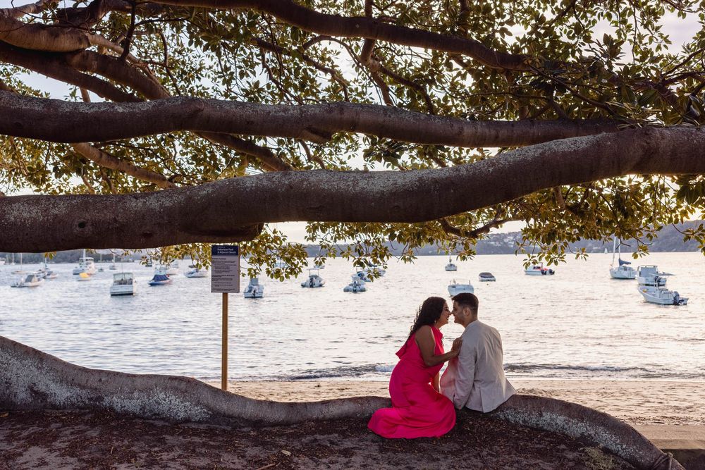 Couple sharing a romantic moment under a large tree by the waterfront with sailboats in the background.
