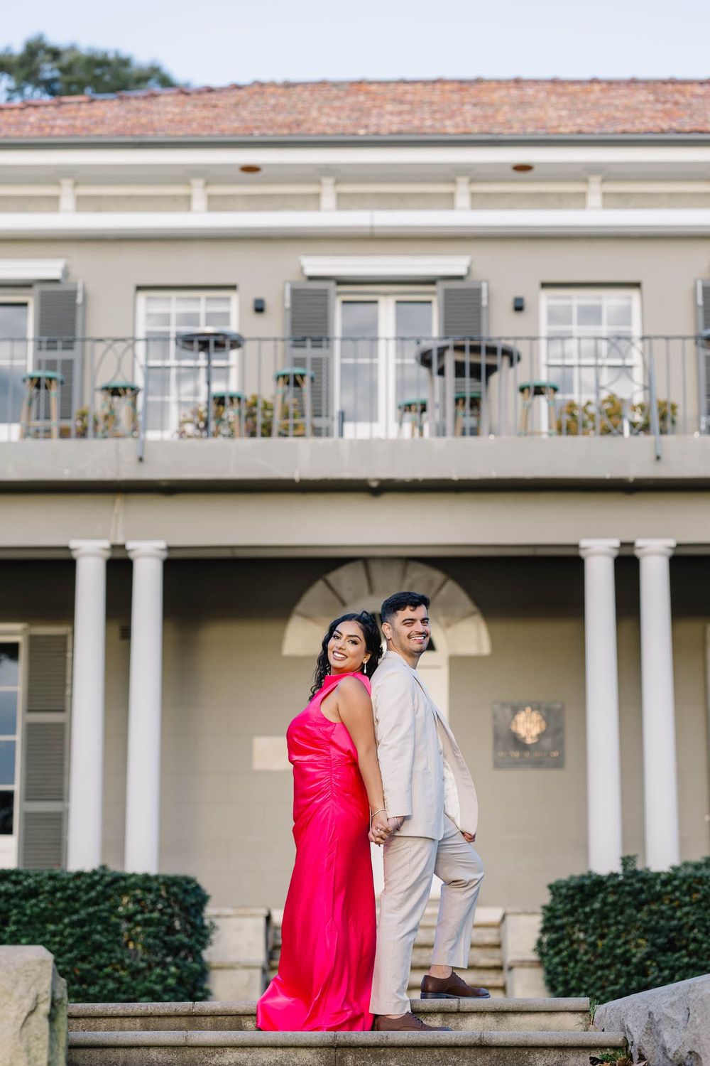 Couple in pink gown and beige suit posing back-to-back on steps before an elegant colonial-style mansion.