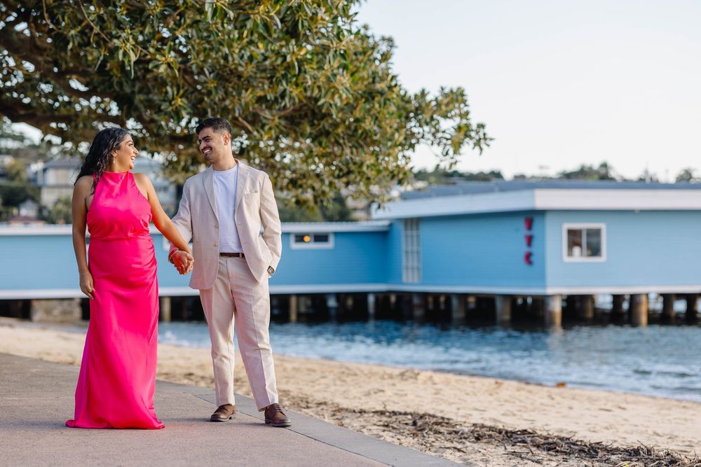 Couple in formal wear holding hands on waterfront path, woman in pink gown, man in beige suit, blue pier behind.