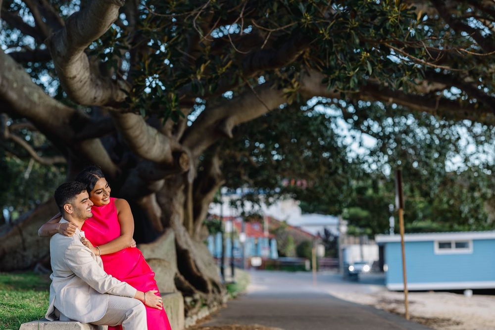 Couple embracing on a bench under a large banyan tree, woman in pink dress, man in white suit, romantic outdoor setting.