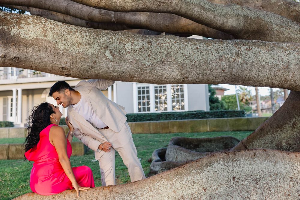 Couple sharing a romantic moment under large banyan tree roots, woman in pink dress, man in beige suit.