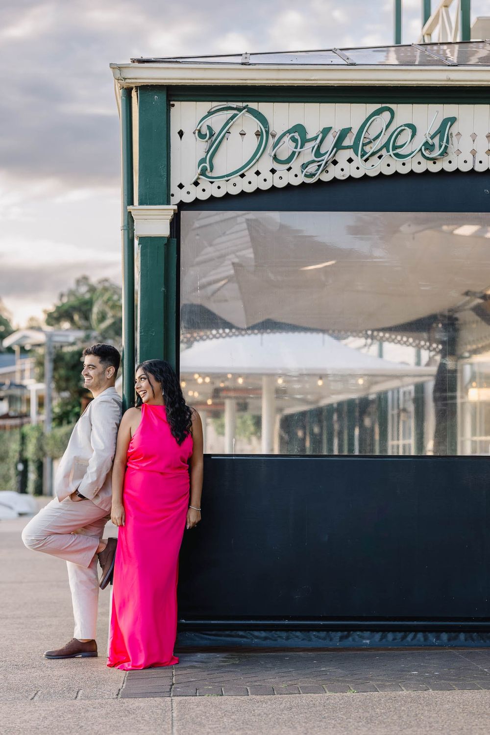 Couple posing outside Doyles restaurant, woman in pink dress, man in white suit, waterfront location.