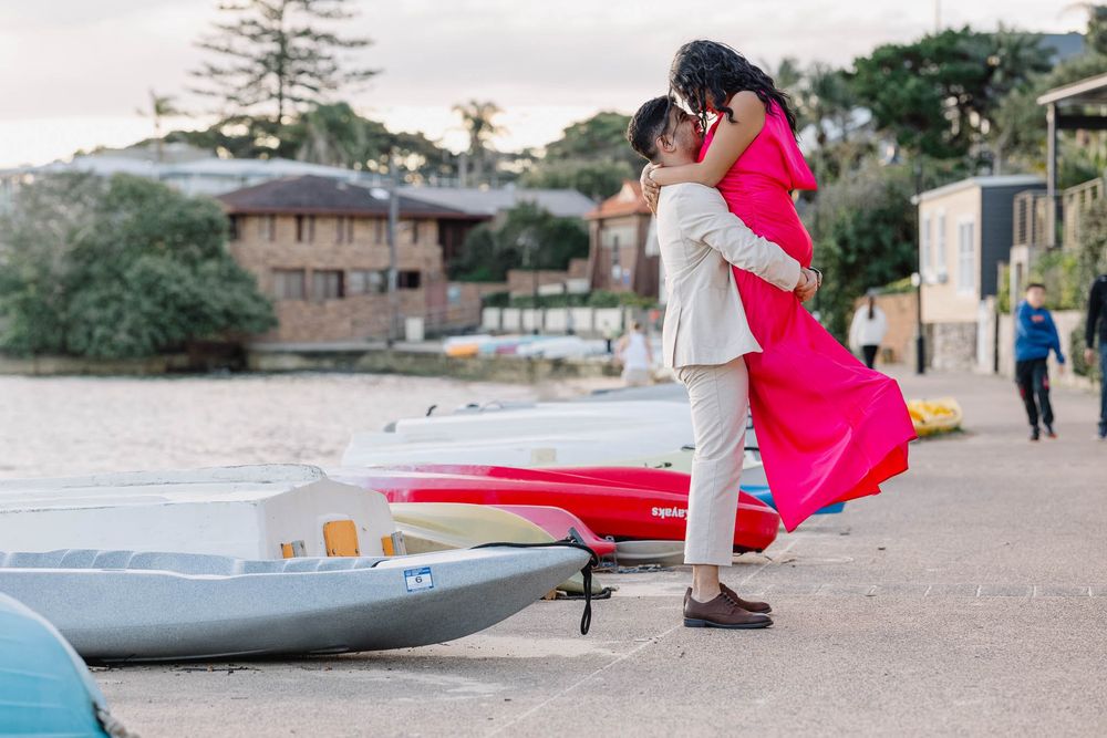 Couple embracing near colorful boats at a waterfront, woman in a vibrant pink dress.