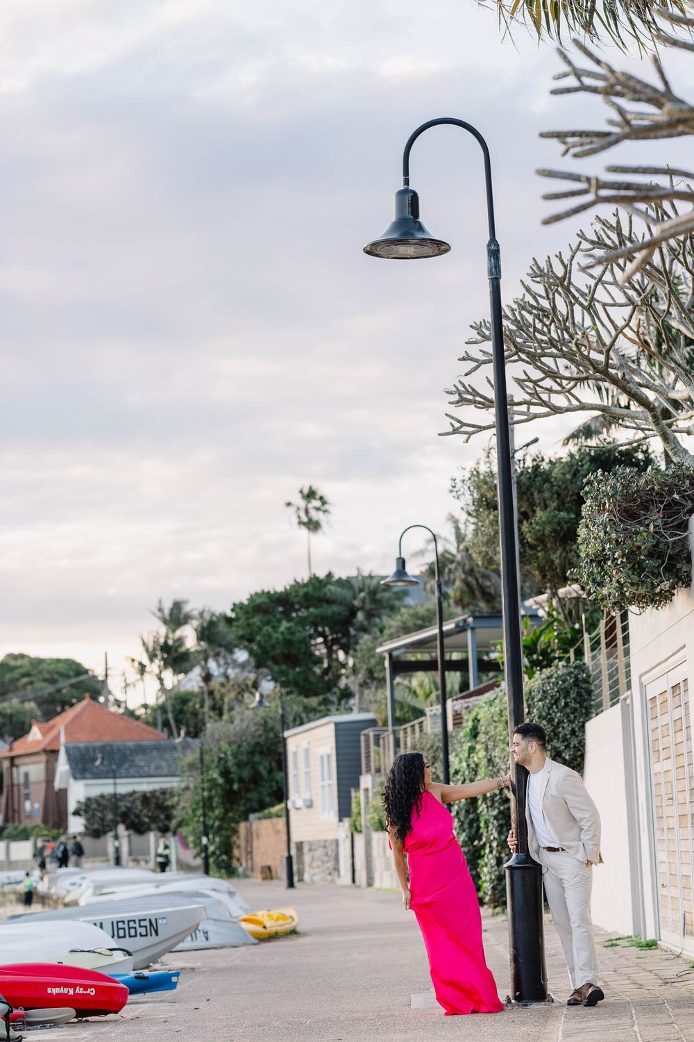 Couple in pink dress and grey suit hold hands by a street lamp near a coastal waterfront at dusk.